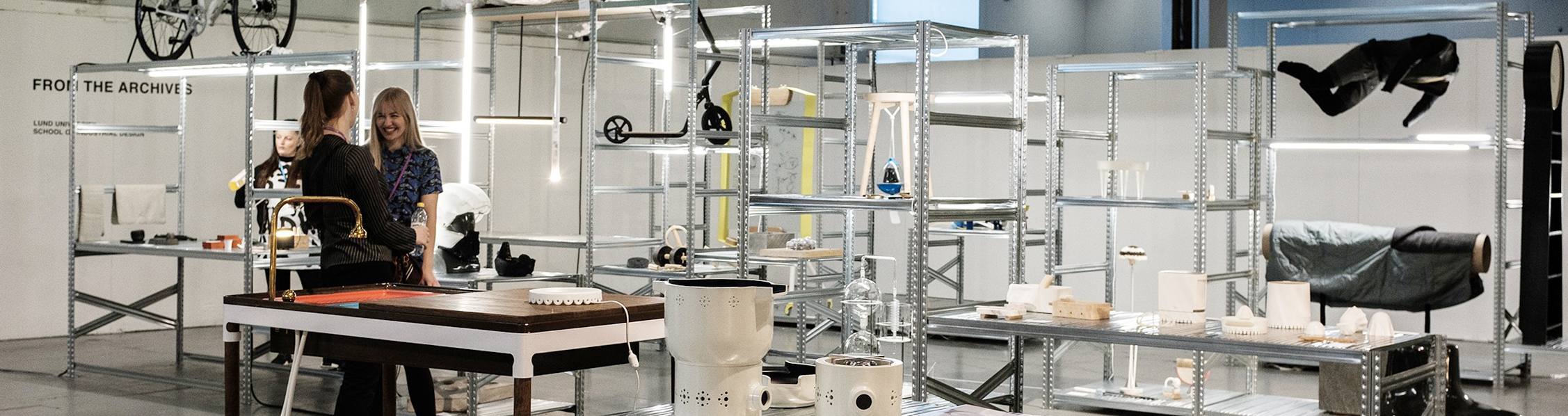 three people standing in a room surrounded by metal shelves and prototypes. Photo. 